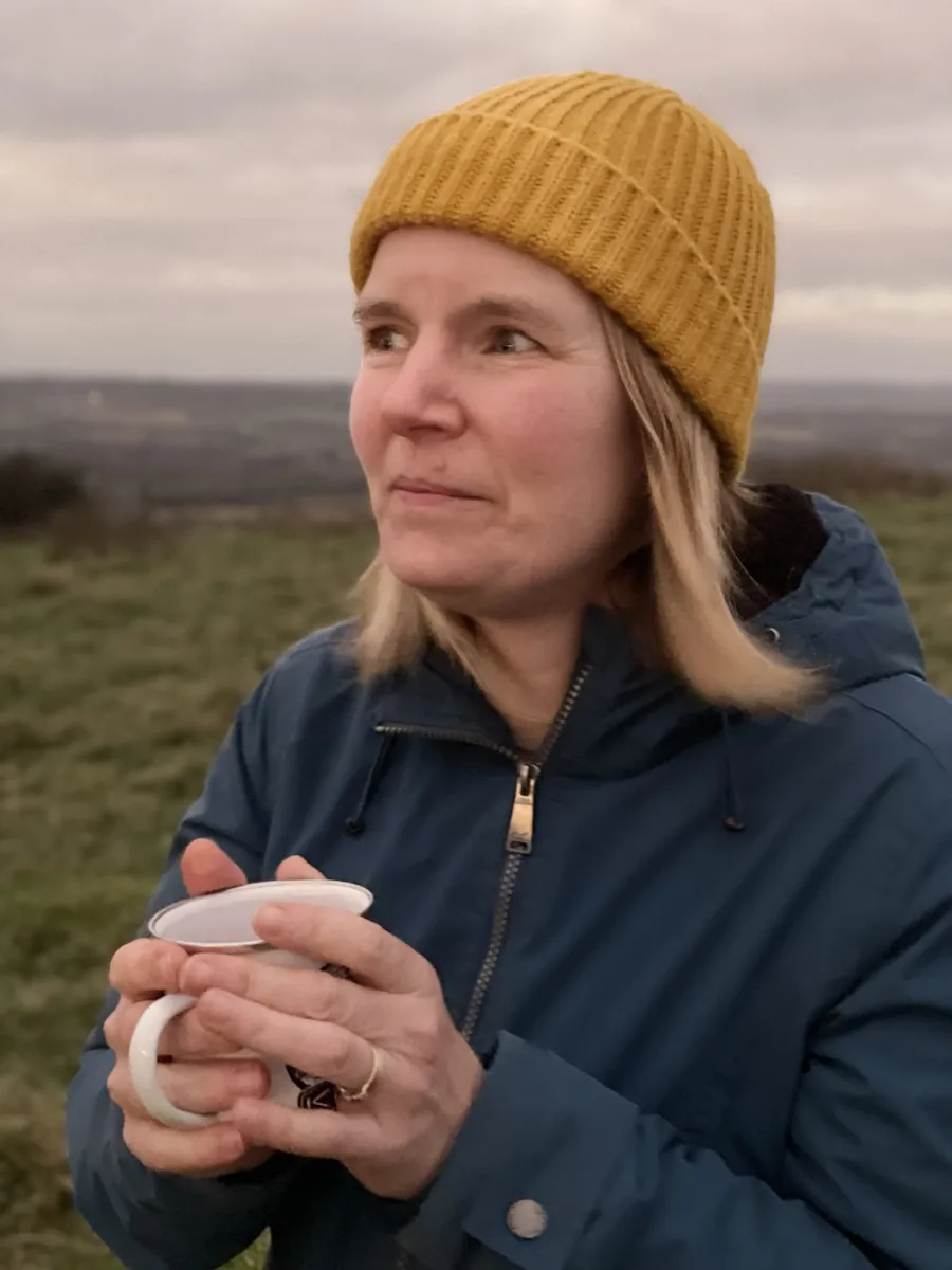 Ruth in a mustard beanie holding a mug of tea on a hilltop